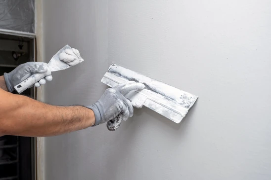 Une main tenant un couteau à enduire applique un enduit de lissage blanc sur un mur intérieur, à la jonction entre une surface grise et blanche